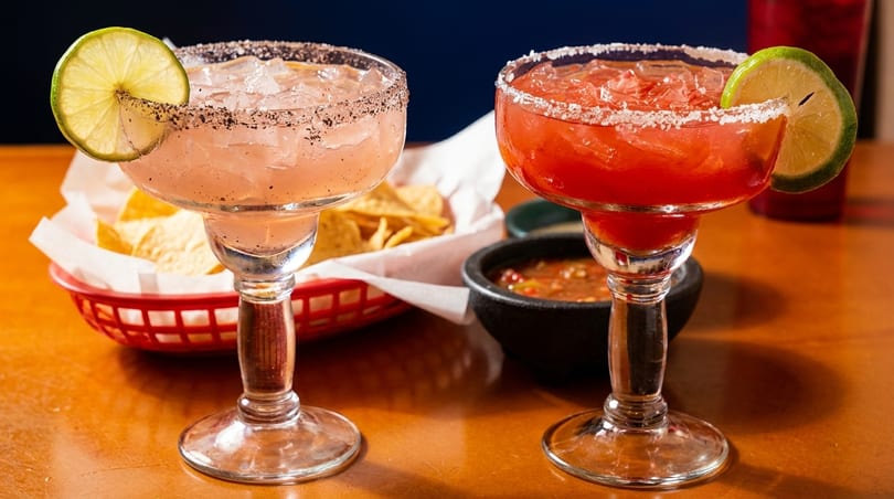 Two margaritas, one pink and one red, with lime slices and salted rims, next to chips and salsa.