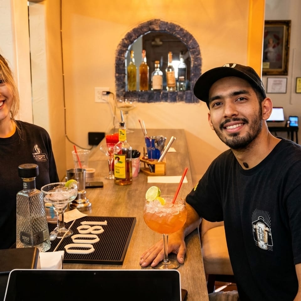 Two smiling El Charro Cafe employees, a woman and a man, stand behind a bar with drinks.
