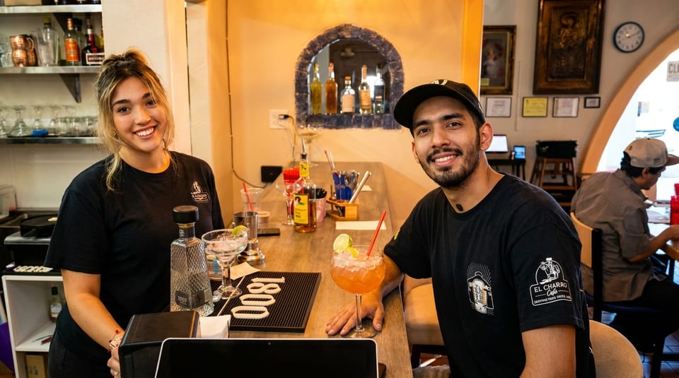 Two smiling El Charro Cafe employees, a woman and a man, stand behind a bar with drinks.