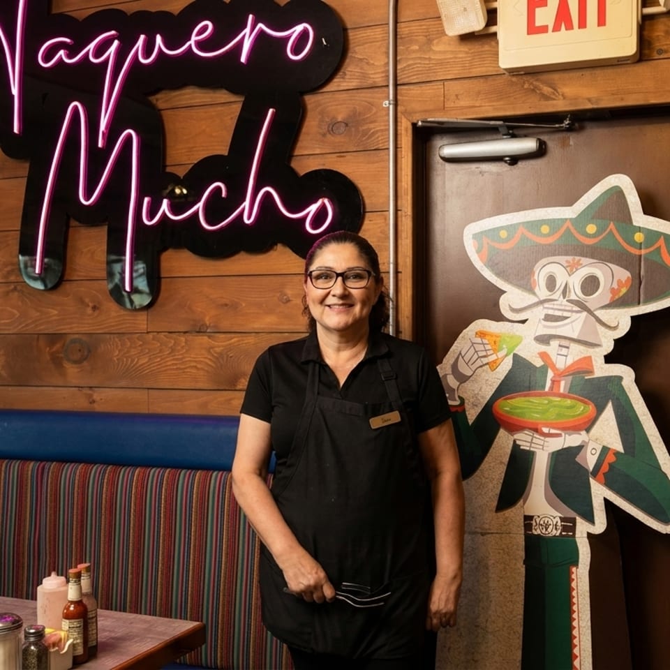 Smiling woman in black apron stands next to a cartoon mariachi cutout and neon sign.
