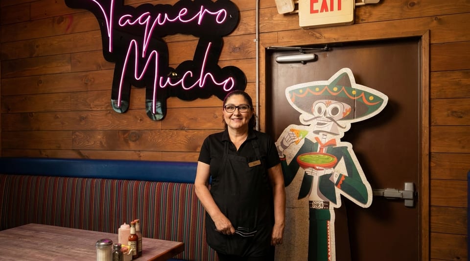 Smiling woman in black apron stands next to a cartoon mariachi cutout and a neon sign.
