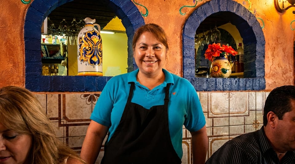 Smiling waitress in a blue shirt and black apron stands in a vibrant Mexican restaurant.