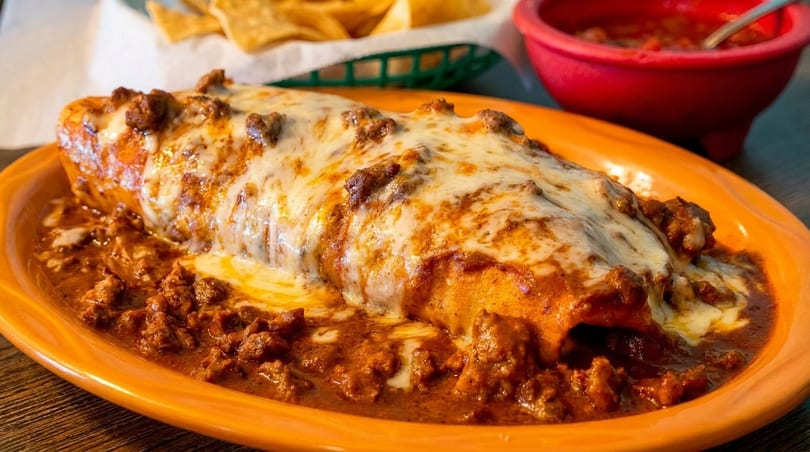 A large, cheesy burrito covered in red sauce and ground beef, served with chips and salsa.