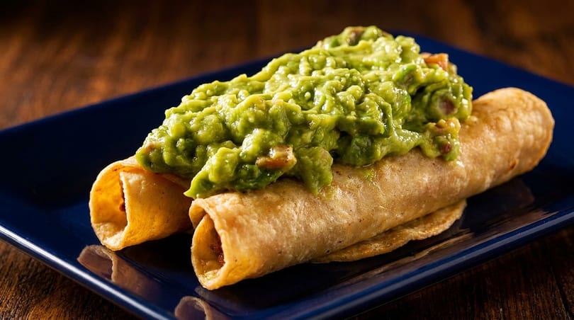 Two golden-brown taquitos topped with a generous serving of chunky guacamole on a dark blue plate.