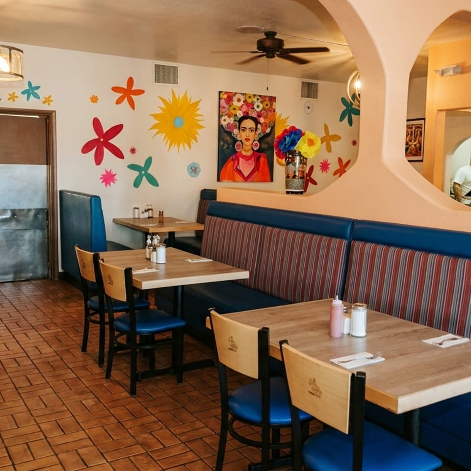 Colorful restaurant interior with blue booths, wooden tables, and a view into the kitchen with chefs.