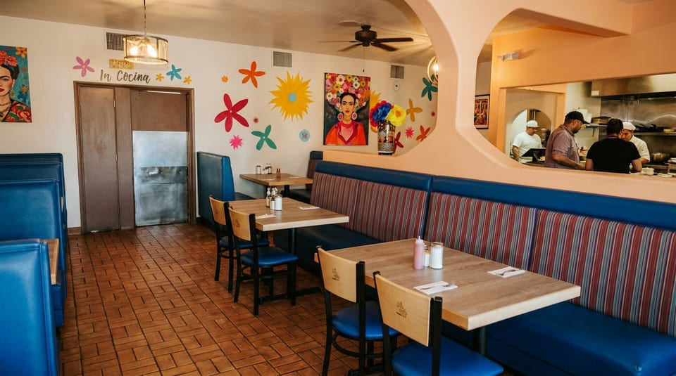 Colorful restaurant interior with blue booths, wooden tables, Frida Kahlo art, and a view into the kitchen.